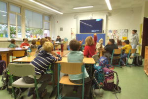 Foto aus dem hinteren Teil einer Klassenzimmers Richtung Tafel. Die Schüler/innen sind von hinten zu sehen wie sie auf ihren Plätzen sitzen. Einige Kinder melden sich.