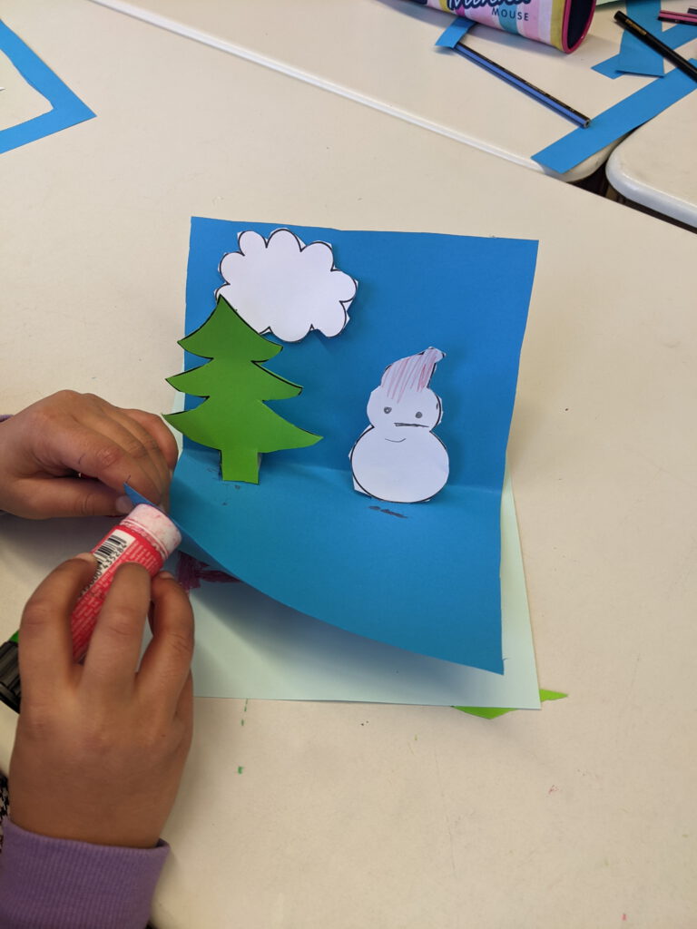 Foto wie eine Weihnachtskarte gebastelt wird. Auf blauem Papier klebt ein SChneemann, eine Wolke udn ein Tannenbaum. Zwei Kinderhände kleben dieses Papier auf ein hellblauen Papier.