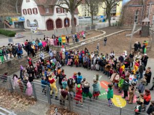Foto verkleideter Grundschulkinder die auf einem Schulhof im großem Halbkreis stehen um einer tanzenden Gruppe zuzusehen.
