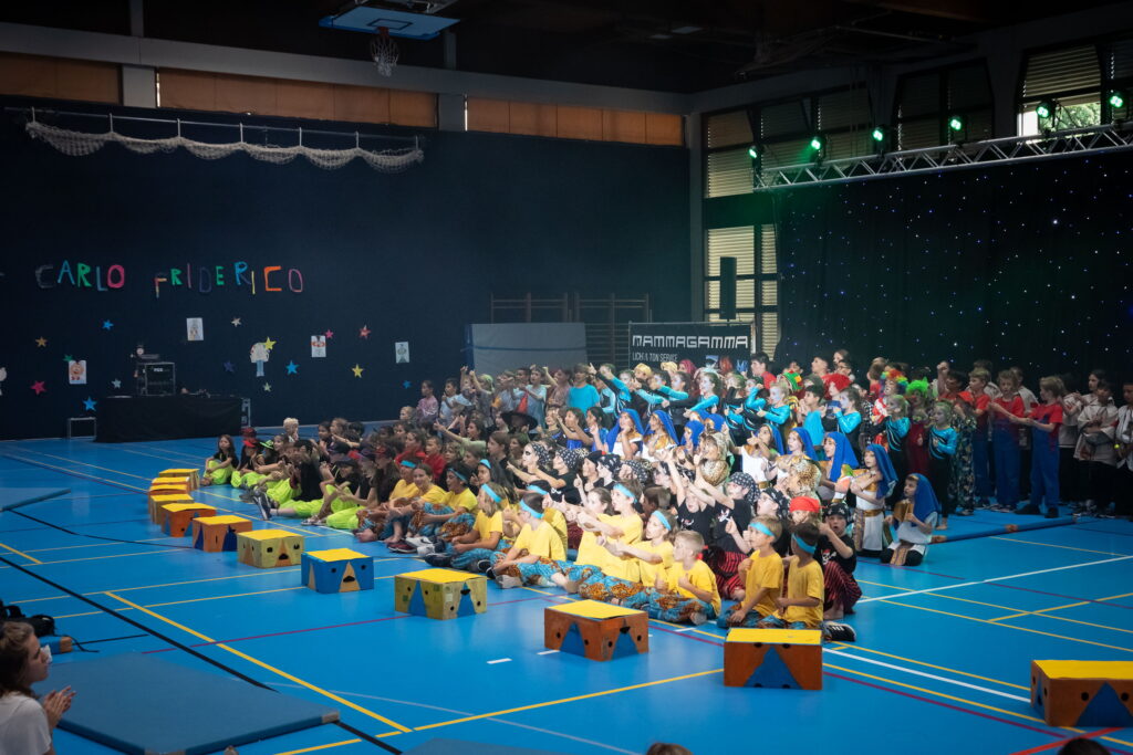 Fotografie einer großen Kindergruppe die in einer zur Manege umgewandelten Sporthalle bunt verkleidet singen und ihre Dauen in die Höhe strecken.
