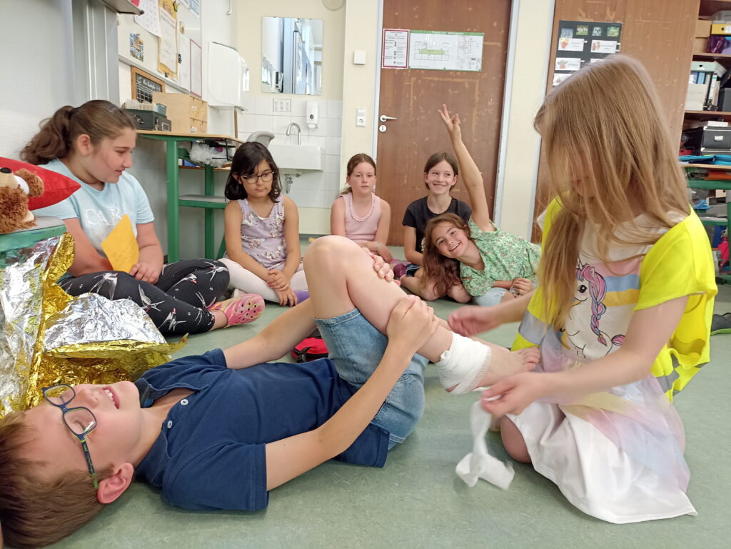Foto einer Gruppe von 7 Kindern, welche im Klassenzimmer auf dem Boden sitzen. Ein Mädchen verbindet einem Jungen, der auf dem Boden liegt und so macht als sei er verletzt, den Knöchel.