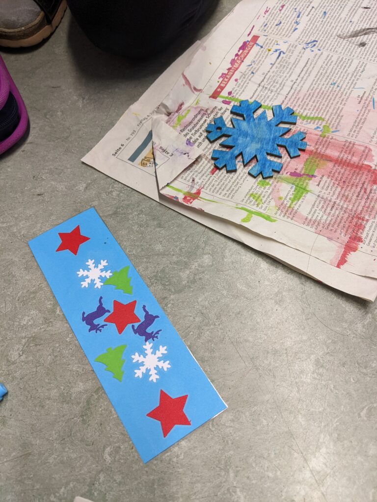 Foto eines selbstgebastelten Lesezeichens aus blauem Papier mit aufgeklebten Sternen, Schneeflocken und Tannenbäumen. Daneben liegt auf einer Zeitung eine Schneeflocke aus Holz welche blau angemalt wurde.
