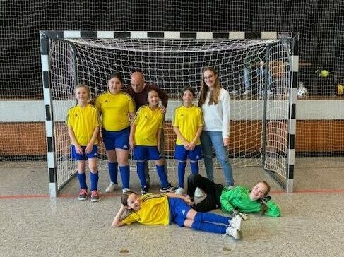 Foto einer Gruppe von 6 Mädchen, einem Mann und zwei die in einem Fußballtor stehen. Zwei der 6 Kinder liegen vor dem Tor.